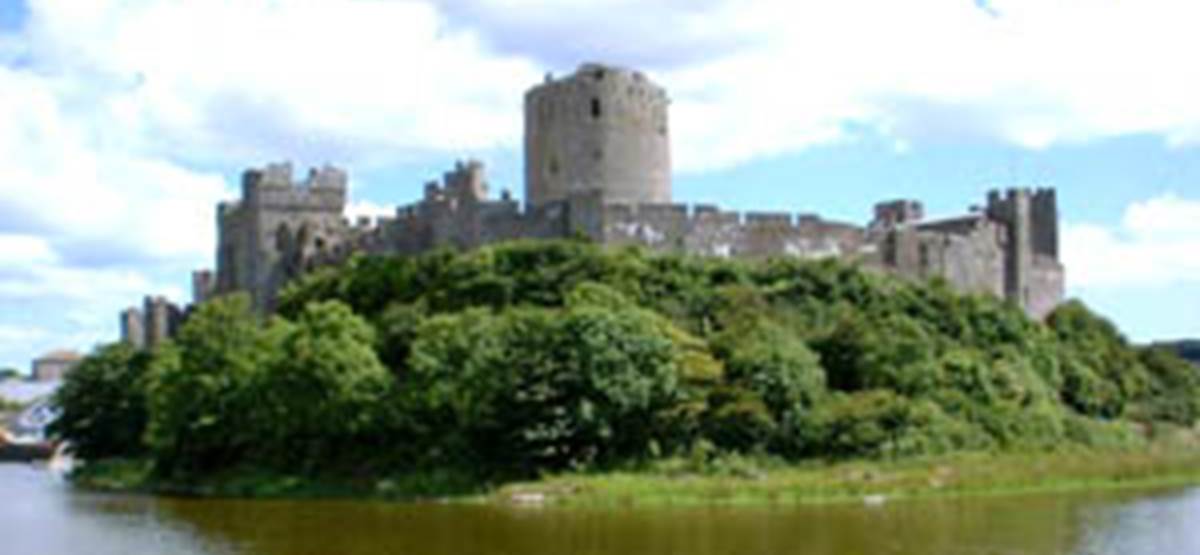 A castle on a hill surrounded by trees with Pembroke Castle in the background

Description automatically generated